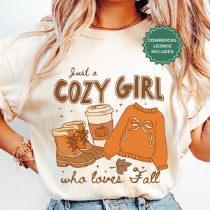 Pode incluir: Uma t-shirt cor de creme com uma estampa gráfica com o texto "Just a COZY GIRL who loves Fall". O design inclui botas de inverno castanhas, um copo de café com um ícone de abóbora, uma camisola laranja com um laço e uma pequena folha.
