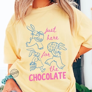 May include: A light yellow t-shirt with a graphic of blue bunnies carrying Easter eggs and a basket. The text "just here for the CHOCOLATE" is in pink. The shirt has a relaxed fit and a round neckline.