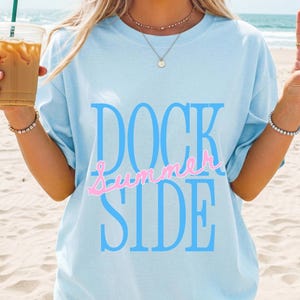 May include: A light blue short-sleeve t-shirt featuring the text DOCK SIDE in large blue letters with the word summer written in a cursive pink script font across the center of the text. The shirt has a crew neckline and a relaxed fit.
