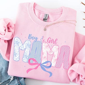 Puede incluir: Una sudadera rosa claro con la palabra "MAMA" en una fuente decorativa, con las letras que presentan patrones florales y a rayas. Las palabras "Boy & Girl" están encima de la palabra "MAMA", y debajo hay un lazo rosa y azul. Un collar y unos vaqueros están al lado de la sudadera.