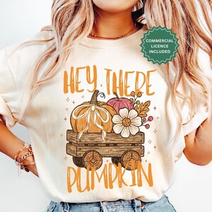Pode incluir: Uma t-shirt cor de creme com um gráfico de uma carroça de madeira carregando uma abóbora com um laço branco, uma abóbora rosa menor e uma flor branca. O texto "HEY THERE PUMPKIN" está impresso em laranja acima e abaixo da ilustração.