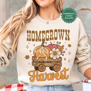 Pode incluir: Uma camisola bege de gola redonda com uma impressão gráfica com o texto HOMEGROWN Harvest. O design apresenta um carrinho de madeira com uma abóbora com um laço branco, uma abóbora rosa mais pequena e uma flor branca, rodeado por estrelas.