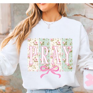 May include: White sweatshirt with a floral print and the word "PRAY" in pink letters. A pink bow is centered below the word.