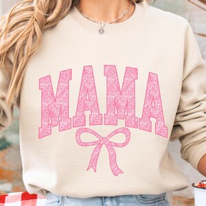 May include: A light beige sweatshirt with a pink "MAMA" graphic with a pink bow underneath. The text is in a decorative font with a white outline.