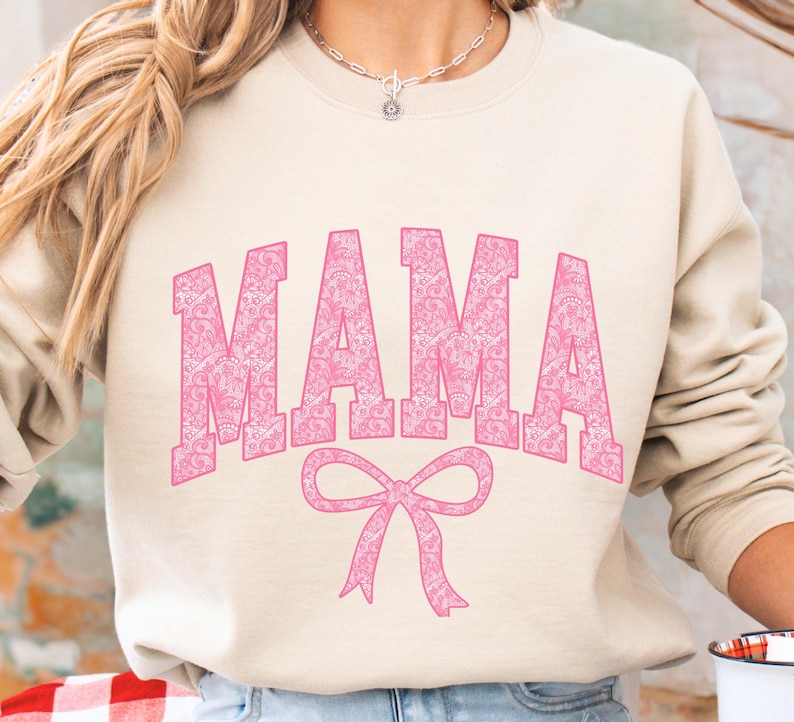 May include: A light beige sweatshirt with a pink "MAMA" graphic with a pink bow underneath. The text is in a decorative font with a white outline.