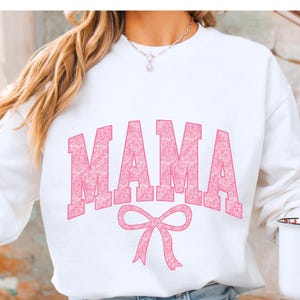 May include: White sweatshirt with a pink "MAMA" graphic in a patterned font with a pink bow underneath.