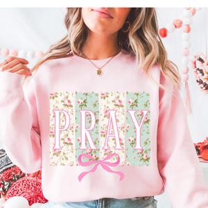 May include: Pink sweatshirt with a floral print and the word "PRAY" in white letters. A pink bow is below the word.