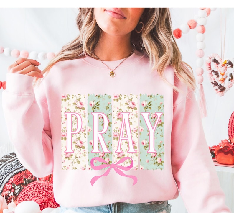 May include: Pink sweatshirt with a floral print and the word "PRAY" in white letters. A pink bow is below the word.