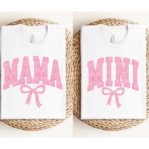May include: Two white t-shirts with pink text. The first shirt says "MAMA" with a pink bow underneath. The second shirt says "MINI" with a pink bow underneath.