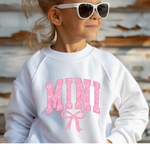 May include: White sweatshirt with a pink "MINI" graphic with a pink bow underneath. The sweatshirt is made of soft, comfortable fabric.