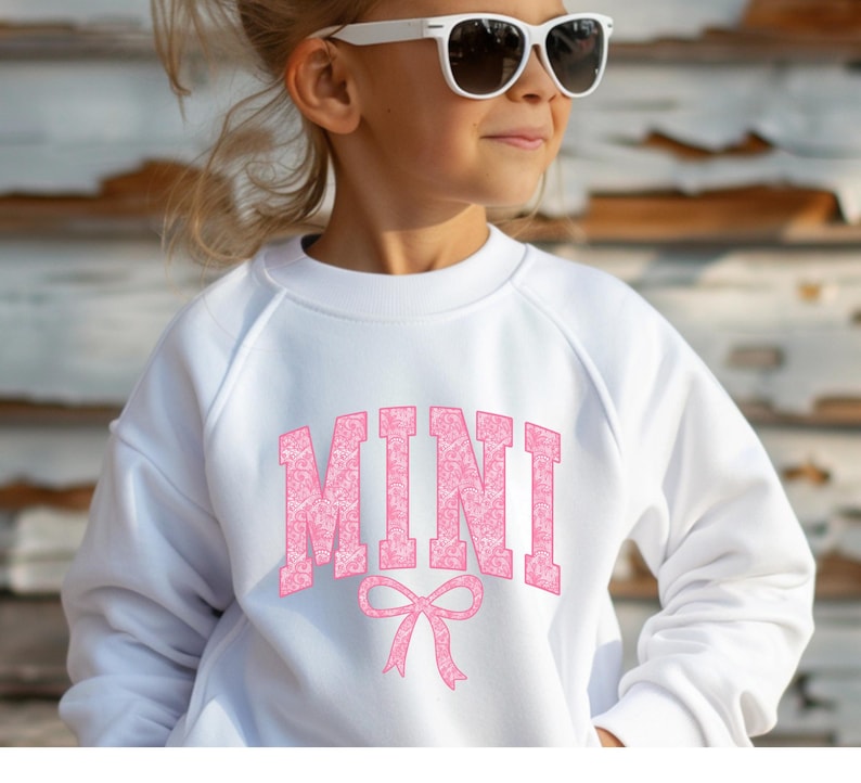 May include: White sweatshirt with a pink "MINI" graphic with a pink bow underneath. The sweatshirt is made of soft, comfortable fabric.