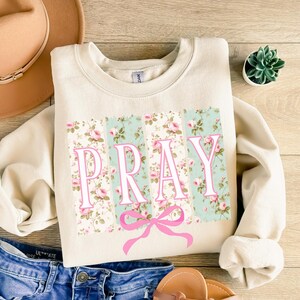 May include: A cream-colored sweatshirt with a floral print and the word "PRAY" in pink letters. A pink bow is centered below the word.