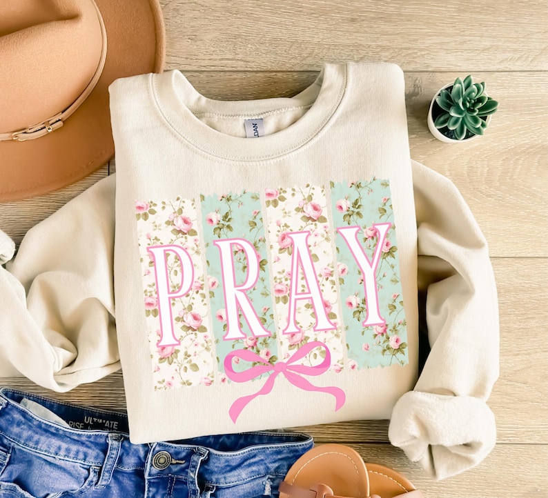 May include: A cream-colored sweatshirt with a floral print and the word "PRAY" in pink letters. A pink bow is centered below the word.