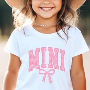 May include: White t-shirt with a pink "MINI" graphic with a pink bow underneath. The graphic has a floral pattern.
