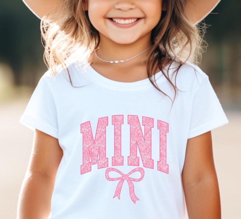 May include: White t-shirt with a pink "MINI" graphic with a pink bow underneath. The graphic has a floral pattern.