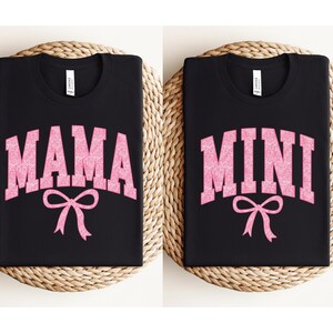 May include: Two black t-shirts with pink glitter lettering. One shirt says "MAMA" with a pink bow and the other says "MINI" with a pink bow.