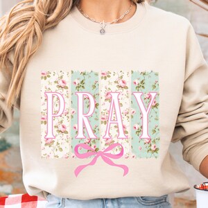 May include: A beige sweatshirt with a floral print design and the word "PRAY" in pink letters. A pink bow is centered below the word.