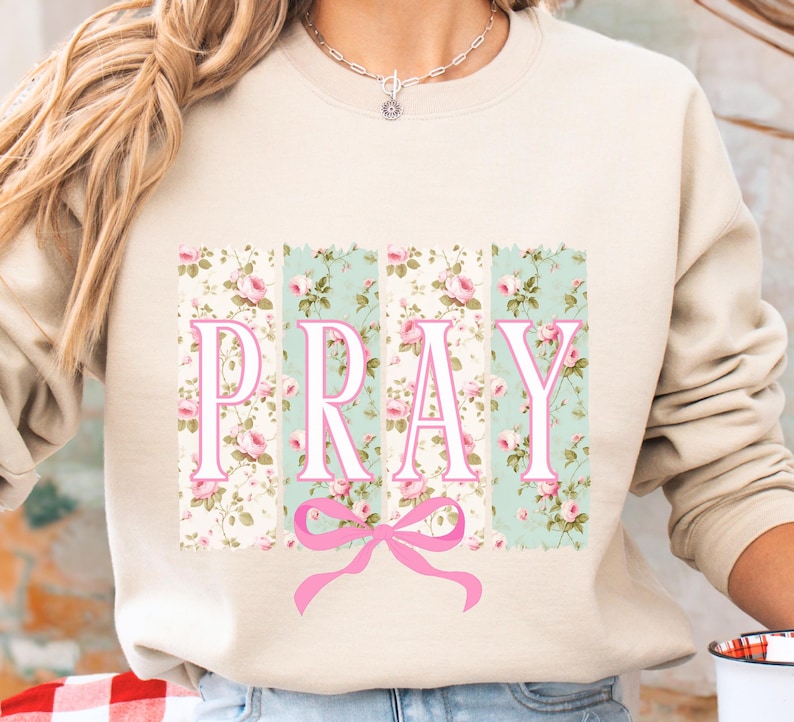 May include: A beige sweatshirt with a floral print design and the word "PRAY" in pink letters. A pink bow is centered below the word.
