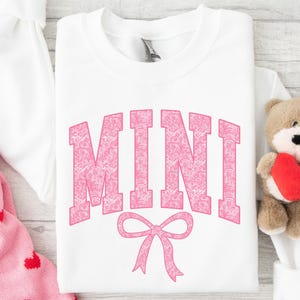 May include: White t-shirt with the word "MINI" in pink with a pink bow underneath. The word "MINI" is made up of a pink floral pattern.