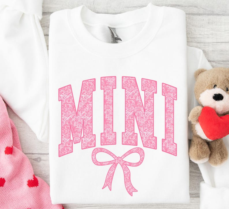 May include: White t-shirt with the word "MINI" in pink with a pink bow underneath. The word "MINI" is made up of a pink floral pattern.
