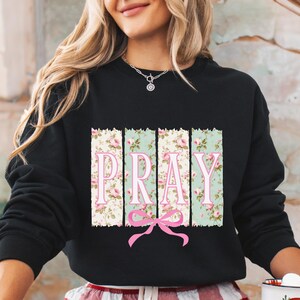 May include: Black sweatshirt with a floral print and the word "PRAY" in pink letters. The floral print is in shades of green, pink, and white.