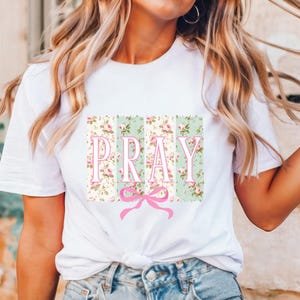 May include: White t-shirt with a floral print design and the word "PRAY" in pink letters. A pink bow is below the word.