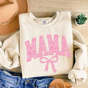 May include: A cream colored sweatshirt with the word "MAMA" in pink with a floral pattern. The word is in a large, bold font and is centered on the sweatshirt. Below the word is a pink bow.