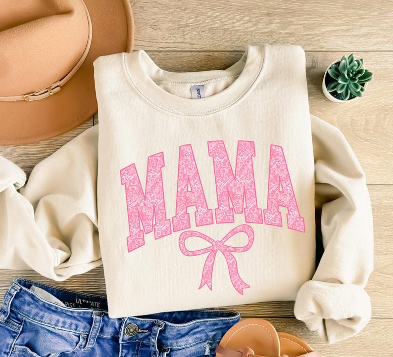 May include: A cream colored sweatshirt with the word "MAMA" in pink with a floral pattern. The word is in a large, bold font and is centered on the sweatshirt. Below the word is a pink bow.