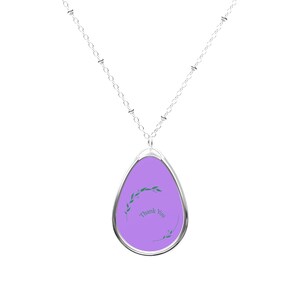 Peut inclure: Un collier en chaîne argent avec un pendentif en forme de larme violette. Le pendentif est orné d'un motif floral vert et des mots "Merci" écrits dessus.