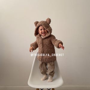 May include: A brown crocheted teddy bear hooded jacket with a white background. The jacket has a zipper closure and is worn by a child standing on a white chair. The text "@thisisolyacrochet" is visible at the bottom of the image.
