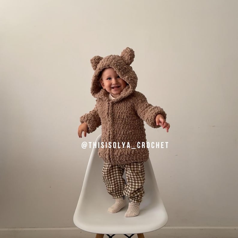 May include: A brown crocheted teddy bear hooded jacket with a white background. The jacket has a zipper closure and is worn by a child standing on a white chair. The text "@thisisolyacrochet" is visible at the bottom of the image.