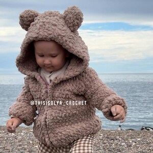May include: A brown crocheted hooded jacket with bear ears. The jacket is worn by a child standing on a beach. The text '@thisisoly_crochet' is visible on the jacket.