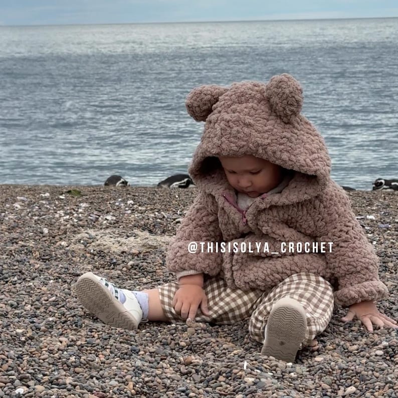 May include: A baby wearing a brown teddy bear hooded jacket with a zipper and plaid pants sits on a beach of small pebbles. The baby is looking down at the pebbles. The text "@thisisolyacrochet" is on the jacket.