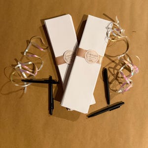 May include: Two stacks of long white rectangular cards wrapped in tan paper bands with circular labels reading Cherished Chapters. Each stack is accompanied by black pens and iridescent curling ribbons arranged around the stationery sets.