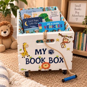 May include: A white wooden storage crate on wheels with the text MY BOOK BOX, decorated with giraffe, monkey, and lion cutouts. The crate contains several children's board books and features a rope handle with a blue grip for pulling.