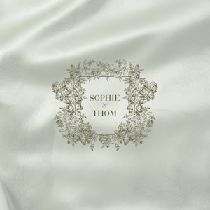 May include: A white satin background with a floral wreath surrounding a shield with the text "Sophie & Thom" in a brown font.