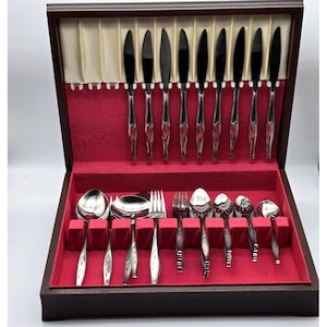 May include: A wooden flatware storage chest with a red velvet interior lining. The set includes eight stainless steel dinner knives held in the lid, and a variety of spoons and forks arranged in the base, all featuring a decorative handle pattern.