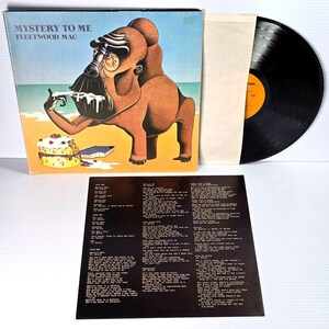 May include: Vintage vinyl record album titled "Mystery to Me" by Fleetwood Mac. The album cover features a cartoon illustration of a brown ape-like figure eating cake on a beach. Includes the record, inner sleeve, and lyric sheet.