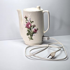 May include: Cream-colored electric coffee pot with a floral rose design. The pot has a ribbed texture, a lid, and a handle. A white power cord and plug are attached. The floral design features pink and purple roses with green leaves.