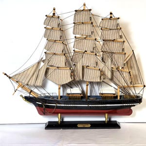 May include: A detailed model of the sailing ship Cutty Sark, featuring beige and white striped sails, black hull, and red waterline. The model is mounted on a black base with gold accents and a nameplate.