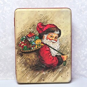 May include: Vintage rectangular tin with a festive illustration of Santa Claus carrying a basket filled with gifts. Santa is depicted in a red suit and hat, with a white beard and a jolly expression. The tin has a beige background with a red border.