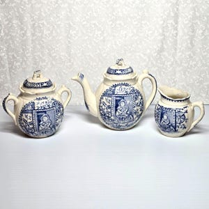 May include: A three-piece antique tea set with a blue and white design. The set includes a teapot, a creamer, and a small teapot. Each piece features a detailed illustration of a person in a window, surrounded by floral patterns.