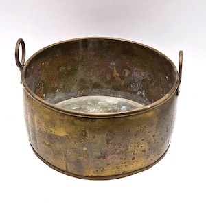 May include: A round, antique brass bowl with two small loop handles. The bowl has a weathered, mottled appearance with shades of gold, brown, and green. The interior shows signs of age and use, adding to its vintage charm.