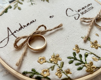 May include: A white fabric ring pillow with embroidered floral designs and the names "Andrea" and "Simona" with the date "11.05.2021". Two gold wedding bands are placed on the pillow, tied with twine.