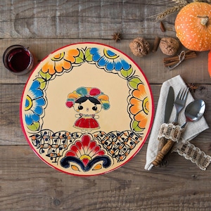 May include: A decorative ceramic plate with a hand-painted design featuring a stylized doll in a red dress and a colorful headdress. The plate has a cream-colored center with a floral border in blue, orange, and green. A red rim surrounds the plate.