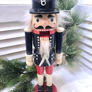 Könnte beinhalten: Traditionelle Holz-Nussknacker-Figur. Sie hat einen schwarzen Zylinder mit weißem Emblem, eine blaue Jacke mit weißen Punkten, rote Shorts und einen weißen Bart. Der Sockel ist rot, vor etwas Grün.