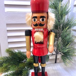 Könnte beinhalten: Ein Holz-Nussknacker in klassischem Design. Die Figur trägt eine rote Jacke, eine schwarze Hose und eine hohe rote Mütze. Das Gesicht ist hellbraun mit einem schwarzen Schnurrbart und weißem Haar. Der Nussknacker steht auf einem roten Sockel.