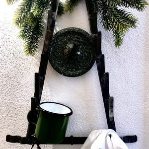 Puede incluir: Una estantería de pared rústica de metal oscuro con una taza esmaltada verde, un plato verde moteado y una toalla blanca con una "S" bordada en verde. La estantería está decorada con ramas de pino artificiales.