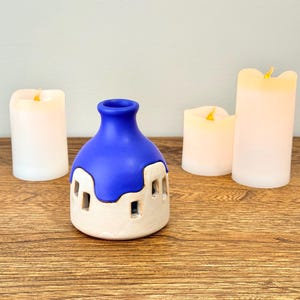 May include: A ceramic candle holder with a blue top and a white base, featuring cut-out windows. The holder is surrounded by three white pillar candles with flickering yellow LED lights, all set on a wooden surface.