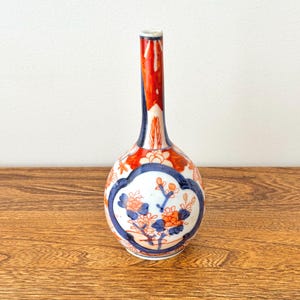 Könnte beinhalten: Kleine, dekorative Keramikvase mit schmalem Hals und rundem Körper. Die Vase hat einen weißen Sockel mit blauen und orangefarbenen Blumendesigns, umrahmt von einem blauen Rand. Der Hals ist rot und weiß, mit glänzendem Finish. Die Vase ist ca. 15 cm hoch.